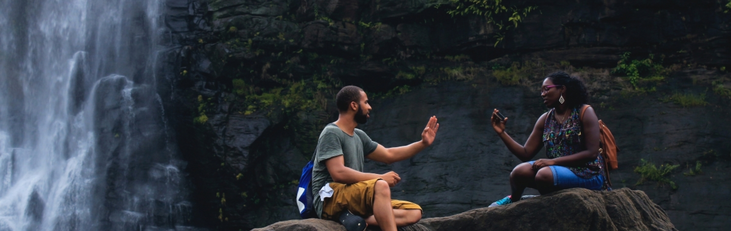 man and woman talking and sitting on a rock near a waterfall