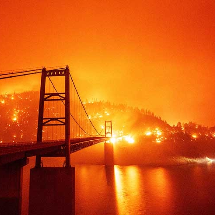 A fire burns on a hill behind a bridge with a bright orange sky